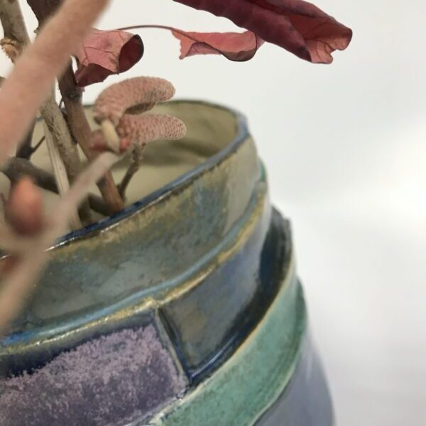 Ręcznie wykonany wazon ceramiczny z kolorowymi, nierównymi pasami w odcieniach błękitu, zieleni i fioletu, o artystycznej, surowej formie.