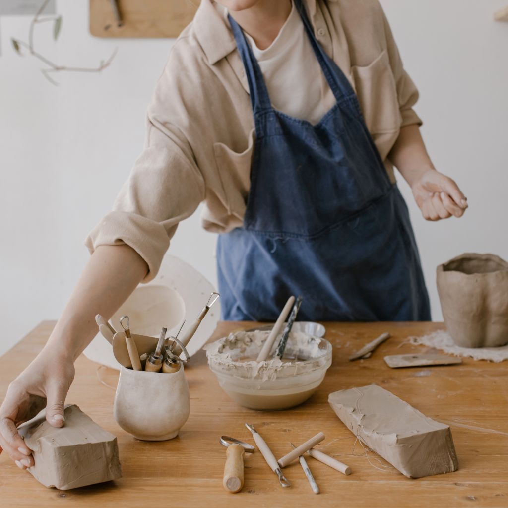 Osoba w fartuchu ceramicznym pracująca z gliną w pracowni. Na stole narzędzia garncarskie, misa z wodą i surowa glina